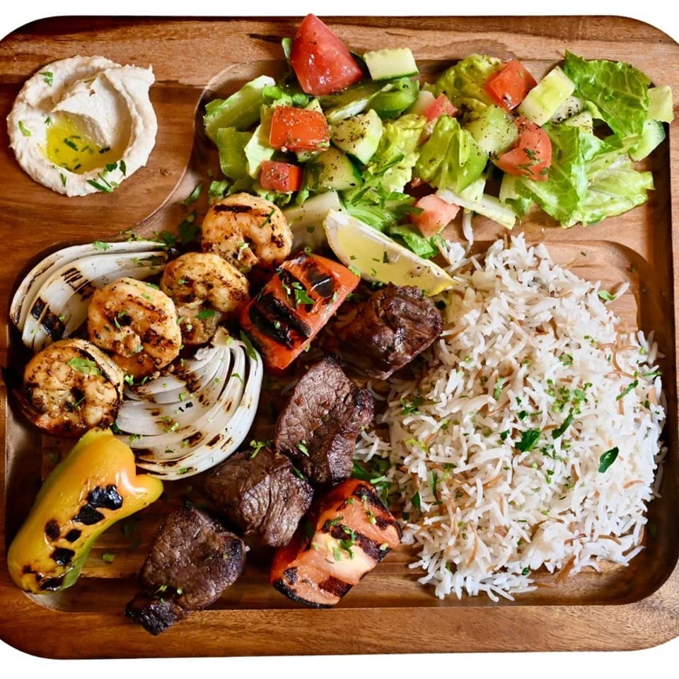 Wooden platter featuring grilled shrimp, beef, vegetables, basmati rice, salad, and hummus.